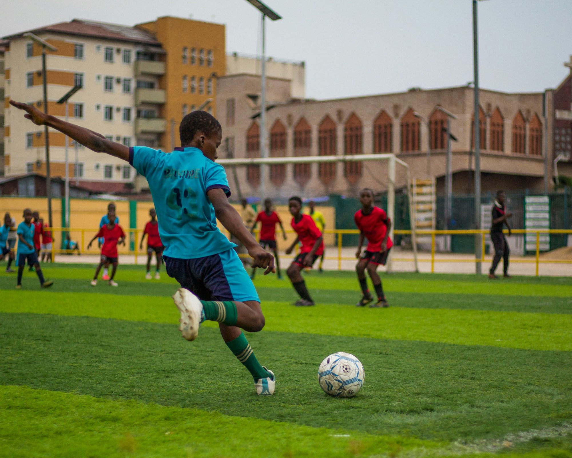 A player striking the ball on a city pitch, opponents and rooftops behind